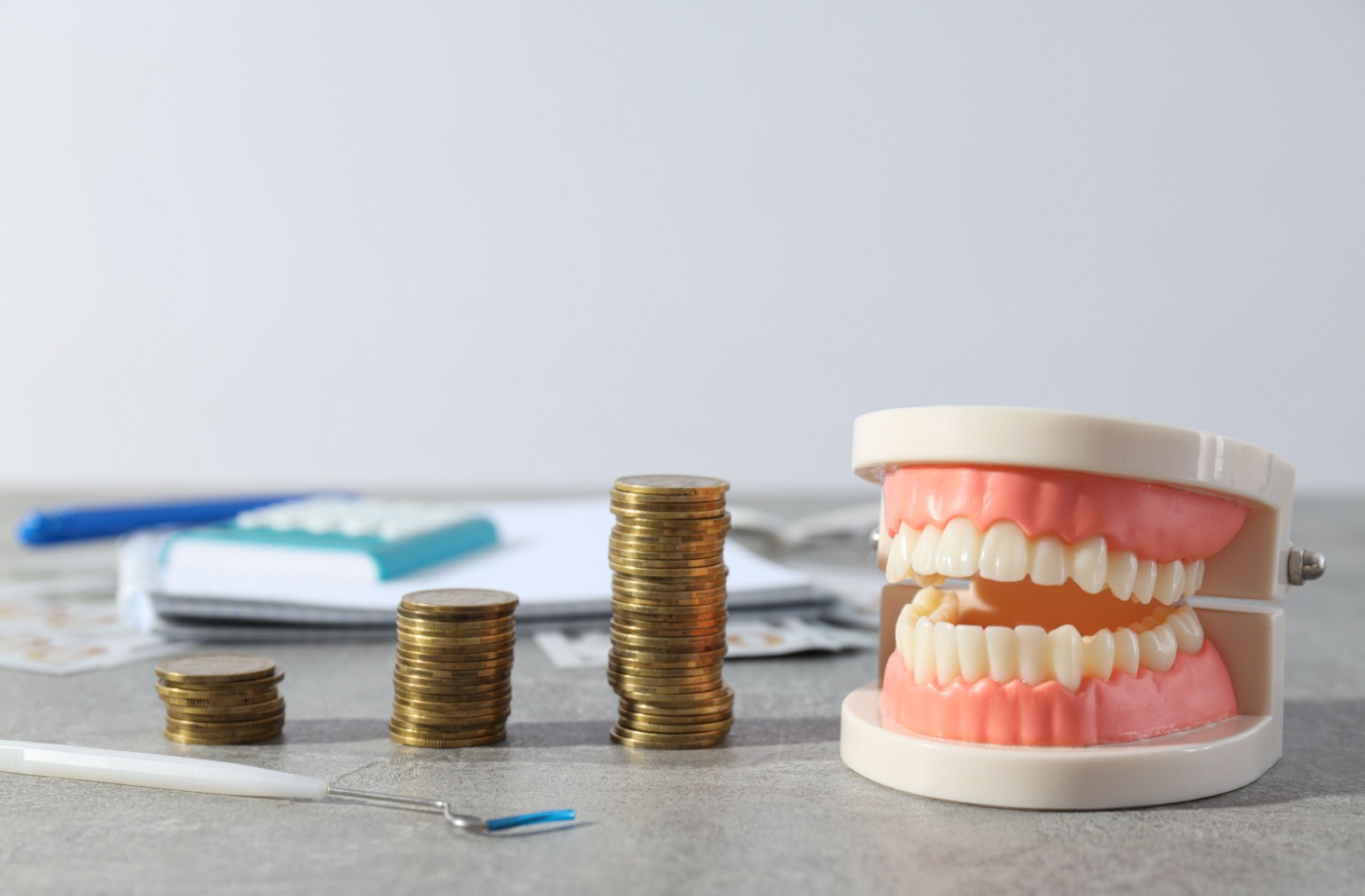 Model teeth sit on a counter next to a dental instrument and stacks of coins with a notebook and calculator in the background.