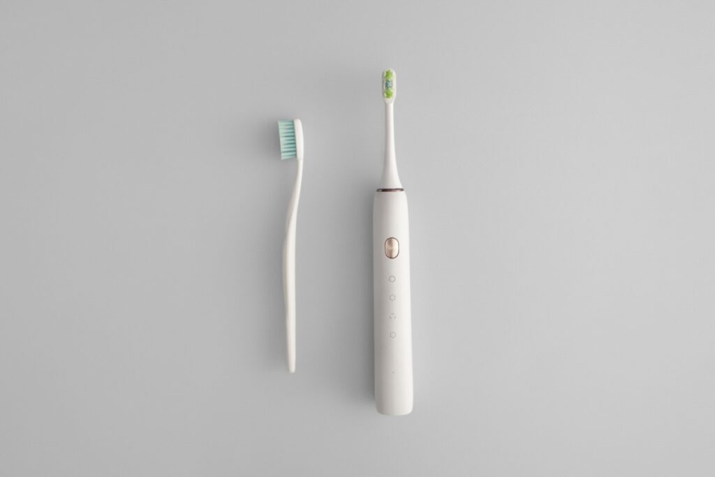 Top-down view of a manual toothbrush beside a white electric toothbrush on a light gray background.