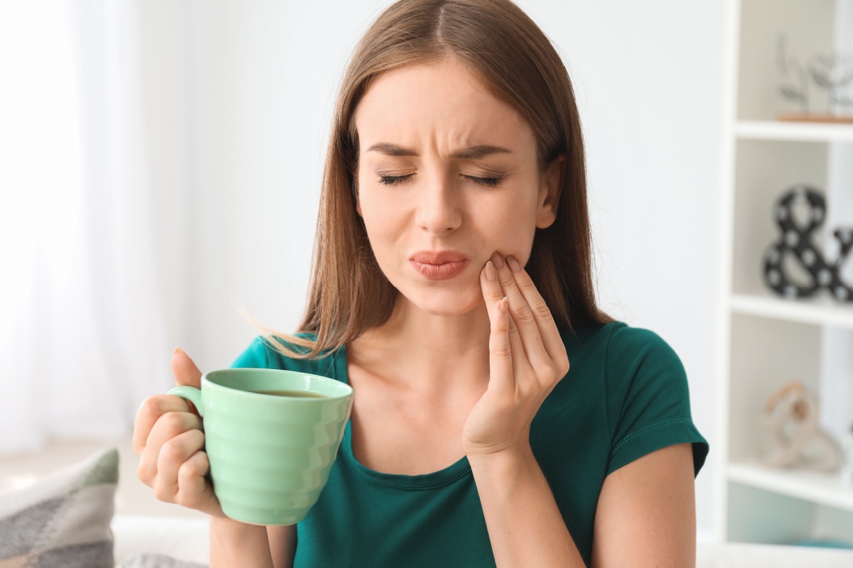 Woman experiencing tooth sensitivity and pain while drinking hot beverage, holding hand to jaw in discomfort at home.